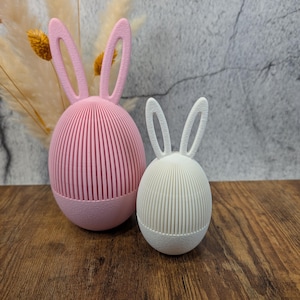 May include: Two Easter bunny ornaments. One is pink, the other is white. Both have a ribbed, egg-shaped body and bunny ears. The ornaments are on a wooden surface, with dried flowers in the background.