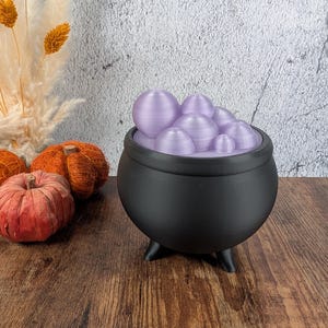 May include: A black cauldron with three legs, filled with lavender-colored orbs. The cauldron sits on a wooden surface, with decorative pumpkins and dried plants in the background. A Halloween-themed decorative item.