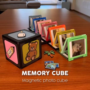 Puede incluir: Un cubo de memoria negro y rosa con una ilustración de oso de peluche y marcos de fotos magnéticos en varios colores. El cubo tiene una ilustración de biberón y etiqueta. El texto "MEMORY CUBE" es visible.