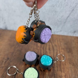 May include: Keyrings in the shape of cauldrons, in shades of orange, purple, turquoise, and green. Each has a black cauldron with brown detailing and a silver chain. The keyrings are shown on a wooden surface.