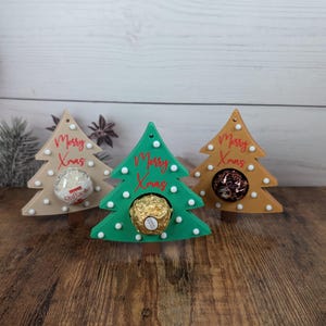 Puede incluir: Tres soportes de madera con forma de árbol de Navidad en beige, verde y dorado. Cada árbol tiene detalles de puntos blancos y el texto rojo "Merry Xmas". Los árboles contienen caramelos envueltos. El fondo es una superficie de madera clara.