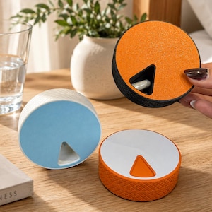 May include: Three circular pill organisers with a rotating lid mechanism. One is orange with a black base, one is light blue with a white base, and one is white with an orange base. Each features a triangular cutout to access the interior compartment.