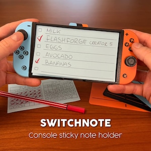 May include: A black plastic sticky note holder shaped like a handheld gaming console with a light blue left controller and an orange right controller. The centre holds a rectangular paper list with handwritten text: MILK, FLASHFORGE CREATOR 5, EGGS, AVOCADO, and BANANAS.