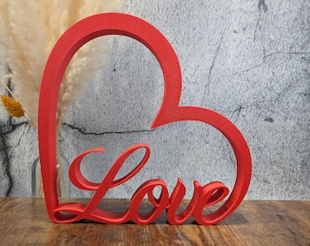 May include: A red, heart-shaped decorative piece with the word "Love" in cursive script. The wooden sign is placed on a wooden surface with a gray textured background. A vase of dried flowers is visible in the background.