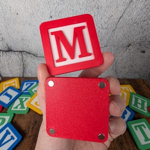Op de afbeelding: Een rode houten blok met de witte letter "M", vastgehouden in een hand. Het blok heeft een rode rand en een witte binnenkant. Andere kleurrijke letterblokken zijn zichtbaar op de achtergrond. De blokken zijn waarschijnlijk voor educatieve doeleinden.