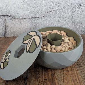 Puede incluir: Un cuenco para frutos secos gris, de forma geométrica, con tapa a juego, lleno de pistachos. La tapa presenta un diseño de hojas en verde y crema. El cuenco tiene un interior verde y un pequeño compartimento central verde oliva.