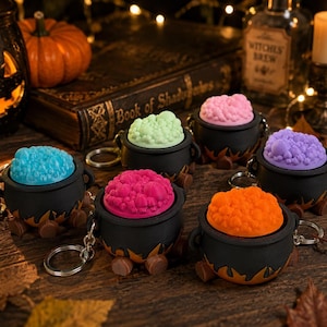 May include: Six black cauldron keychains with textured, bubbling liquid in bright blue, light green, pink, purple, magenta, and orange. Each miniature pot features three small brown feet and a silver metal keyring attached to the side.