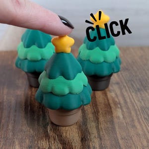 May include: Three miniature Christmas trees with a yellow star on top, each in a brown pot. The trees are shades of green. The word "CLICK" is in the image. A finger is touching one of the trees.