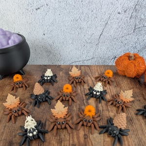 Pode incluir: Uma coleção de decorações de aranhas com tema de Halloween. As aranhas são castanhas e pretas com elementos decorativos como abóboras laranja, folhas bege e caveiras brancas. Uma panela preta com bolhas roxas e uma abóbora de fio laranja também são visíveis.
