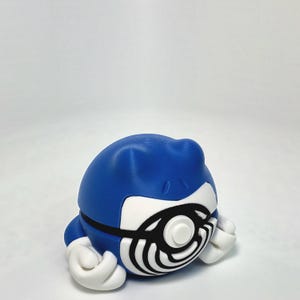 May include: A blue, spherical character figurine with a white face featuring a prominent black spiral pattern. The figure has two small blue ears on top and two white, rounded hands positioned at the base on either side of the central spiral.