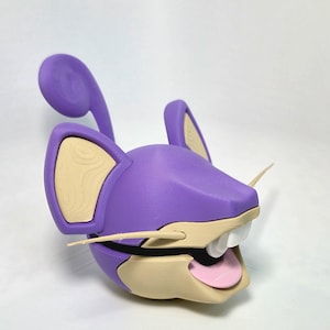 May include: A 3D-printed purple and tan rat head with large ears and whiskers. The rat's mouth is open, revealing white teeth and a pink tongue. The ears have a tan interior with a wood grain pattern.