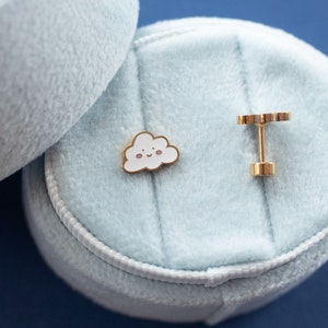 May include: Two gold-colored jewelry pieces in a light blue velvet box. One piece is a small cloud with a smiling face, the other is a simple stud.