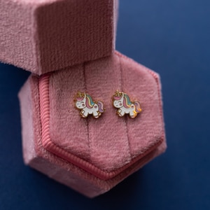 May include: A pair of unicorn stud earrings in a pink velvet jewelry box. The earrings are gold-toned with a white unicorn design and rainbow accents.