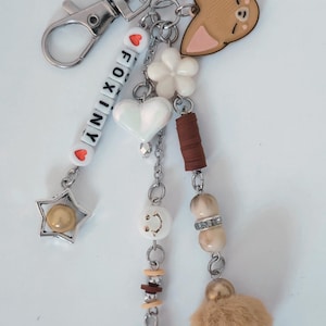 May include: A decorative keychain with various charms. It includes a silver clasp, beads spelling "FOXINY", a star, a heart, a flower, a fox-shaped charm, and a fluffy ball. The colour scheme is silver, white, brown, and beige.
