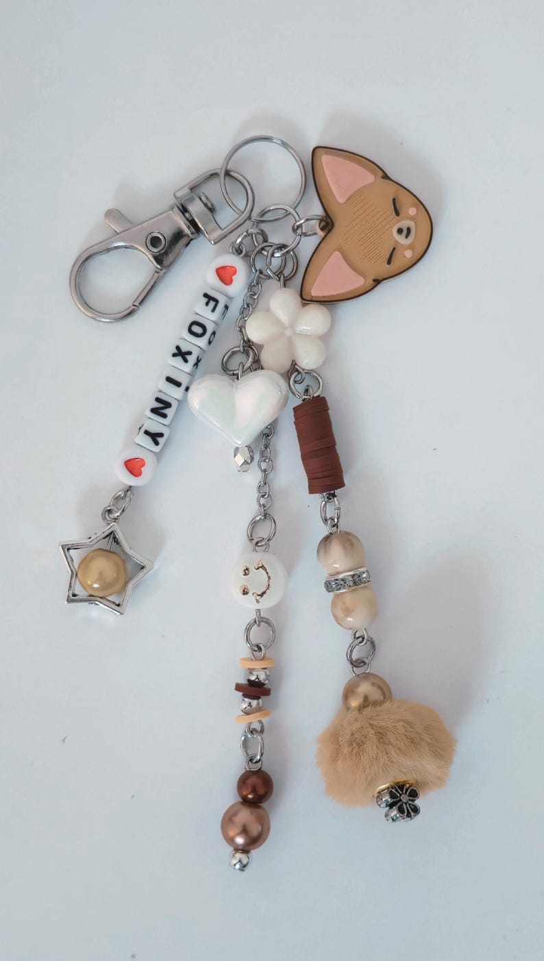 May include: A decorative keychain with various charms. It includes a silver clasp, beads spelling "FOXINY", a star, a heart, a flower, a fox-shaped charm, and a fluffy ball. The colour scheme is silver, white, brown, and beige.