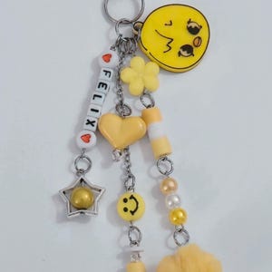 May include: A yellow keychain with various charms, including a star, heart, flower, and a smiley face. The keychain also features the name "FELIX" spelled out with letter beads and a fluffy yellow pom-pom. The metal hardware is silver.