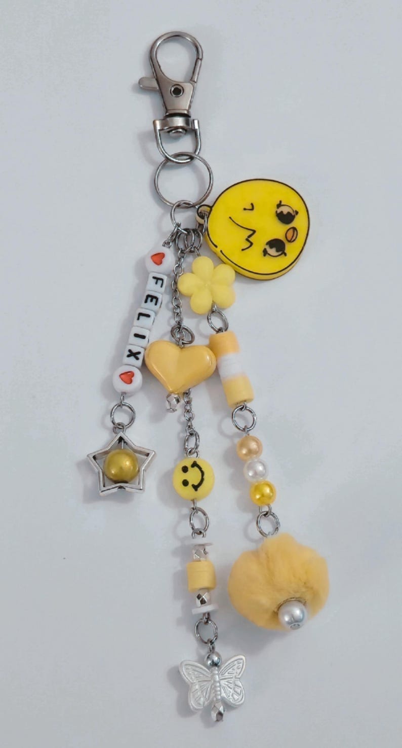May include: A yellow keychain with various charms, including a star, heart, flower, and a smiley face. The keychain also features the name "FELIX" spelled out with letter beads and a fluffy yellow pom-pom. The metal hardware is silver.