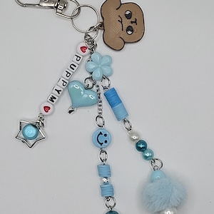 May include: A silver keyring with a cartoon puppy charm and beaded details. The keyring has the word "PUPPY" spelled out with white letter beads, a blue heart, and a star charm. Additional charms include a blue flower, a smiley face, and a fluffy blue pom-pom.
