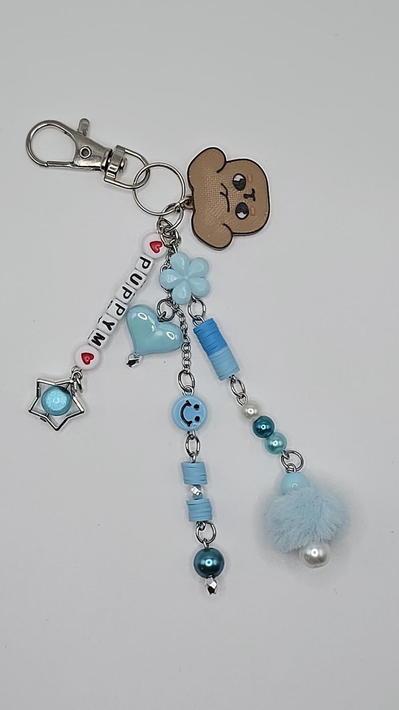 May include: A silver keyring with a cartoon puppy charm and beaded details. The keyring has the word "PUPPY" spelled out with white letter beads, a blue heart, and a star charm. Additional charms include a blue flower, a smiley face, and a fluffy blue pom-pom.