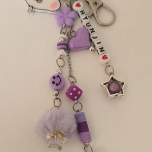 May include: A purple and silver keyring with multiple charms, including a smiley face, a heart, a star, and a fluffy pom-pom. The keyring has the name "HYUNJIN" spelled out in white beads.