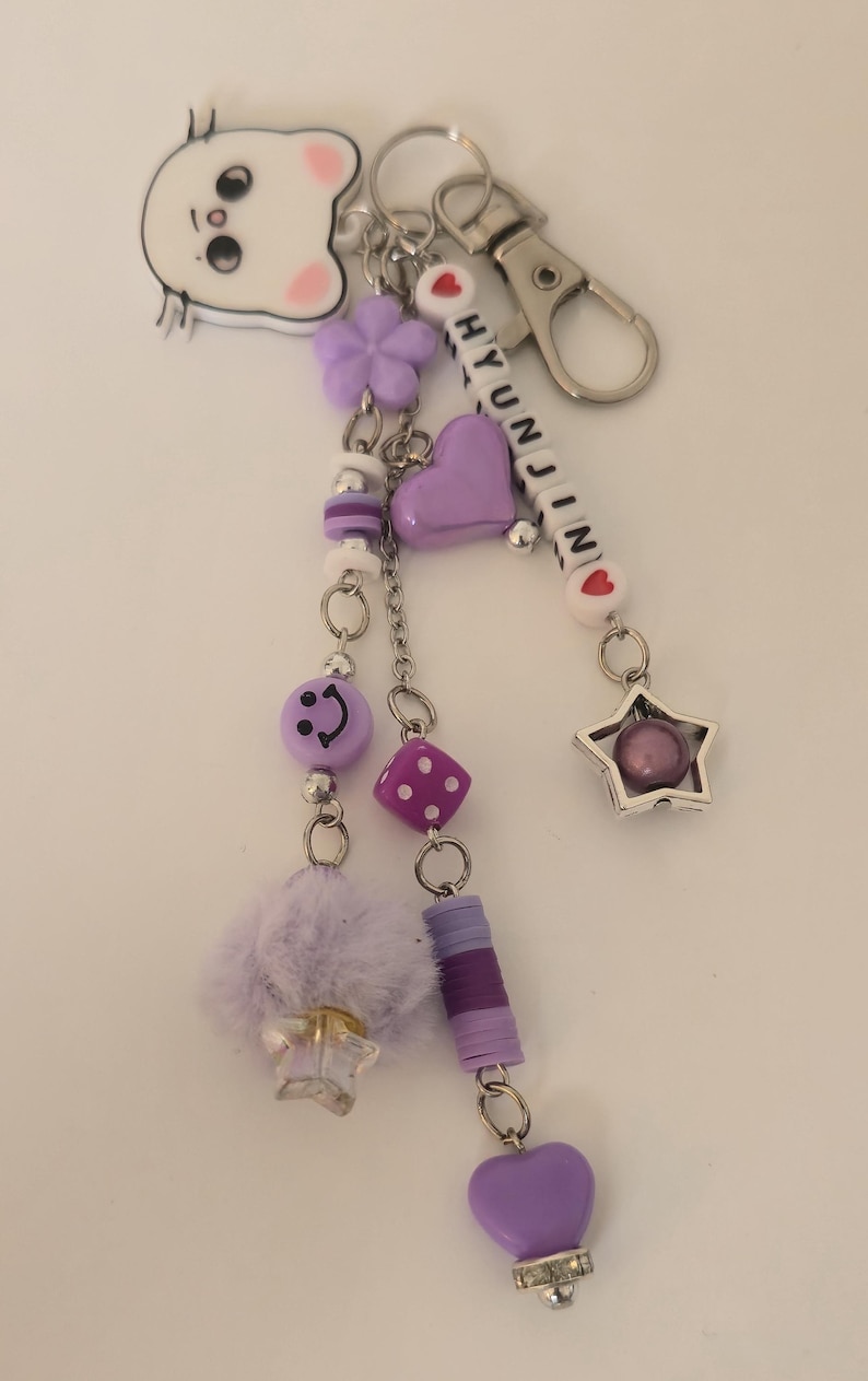 May include: A purple and silver keyring with multiple charms, including a smiley face, a heart, a star, and a fluffy pom-pom. The keyring has the name "HYUNJIN" spelled out in white beads.
