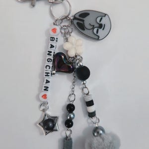 May include: A silver keyring with multiple charms. The charms include a grey cat face, a white flower, a heart, beads, and a star. The keyring also has the name "BANG CHAN" spelled out with letter beads.