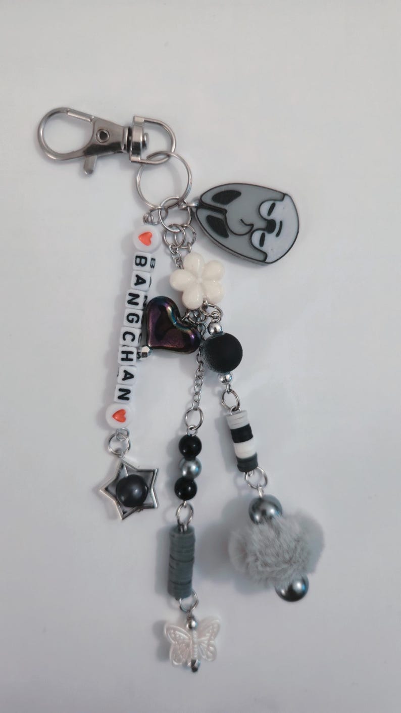 May include: A silver keyring with multiple charms. The charms include a grey cat face, a white flower, a heart, beads, and a star. The keyring also has the name "BANG CHAN" spelled out with letter beads.
