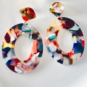 May include: A pair of dangle earrings featuring large oval-shaped pendants with a multicoloured tortoiseshell pattern. The earrings include a smaller, semi-circular top piece connected to the main oval by a small metal ring, showcasing shades of red, blue, white, and orange.