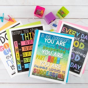 May include: Five colorful posters with inspirational messages for a classroom. The posters have a variety of fonts and colors, including blue, green, yellow, pink, and black. The posters are on a white wooden surface with colorful paper clips.