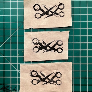 Può includere: Tre toppe di tessuto rettangolari con un disegno di forbici nere. Le forbici sono raffigurate in uno stile grafico audace, con le lame incrociate. Il tessuto è di colore bianco sporco naturale, su uno sfondo verde.