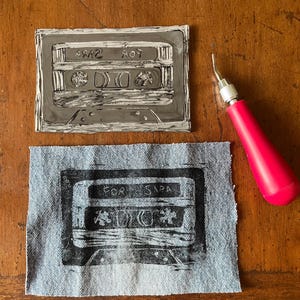 May include: A gray linoleum block print of a cassette tape with the text "FOR SARA" and a matching print on denim. A pink handled carving tool is also in the image. The cassette tape design is a retro theme.