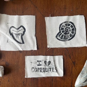 May include: Three small, off-white fabric squares with black linocut prints of a shark tooth, a spiral fossil, and the text "I <3 COPROLITES". The items are arranged on a wooden surface with other fossils.