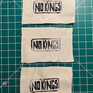 May include: Three rectangular fabric patches with the words "NO KINGS" printed in black block letters. The patches are made of a light beige fabric and are arranged vertically. The background is a green cutting mat.