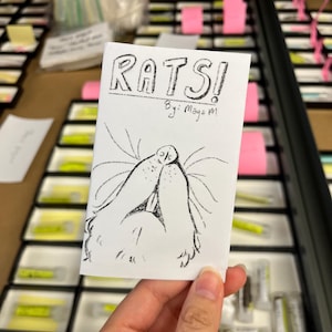 May include: A small, white booklet titled "RATS!" with a hand-drawn illustration of a rat's face. The text "By: Maya M" is below the title. The booklet is held in a hand, with a background of trays and pink sticky notes.