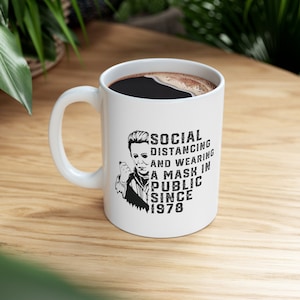 May include: White ceramic coffee mug with a black and white image of a masked figure and the text "SOCIAL DISTANCING AND WEARING A MASK IN PUBLIC SINCE 1978".