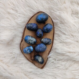Op de afbeelding: Een verzameling van tien gladde, gepolijste, blauwe en witte lapis lazuli stenen, gerangschikt op een houten dienblad. De stenen zijn ovaalvormig en hebben een natuurlijke, organische uitstraling.