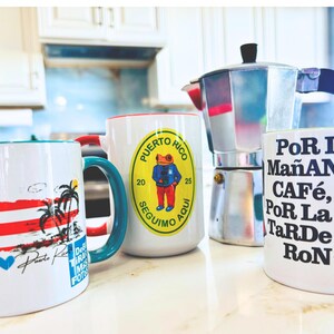 May include: Four white ceramic mugs with different designs. One mug features a Puerto Rican flag, another has a frog wearing a blue shirt, a third has the text "Por La Ma&ntilde;ana Caf&eacute;, Por La Tarde Ron", and the last mug has a palm tree and a beach scene. A silver coffee pot is also in the image.