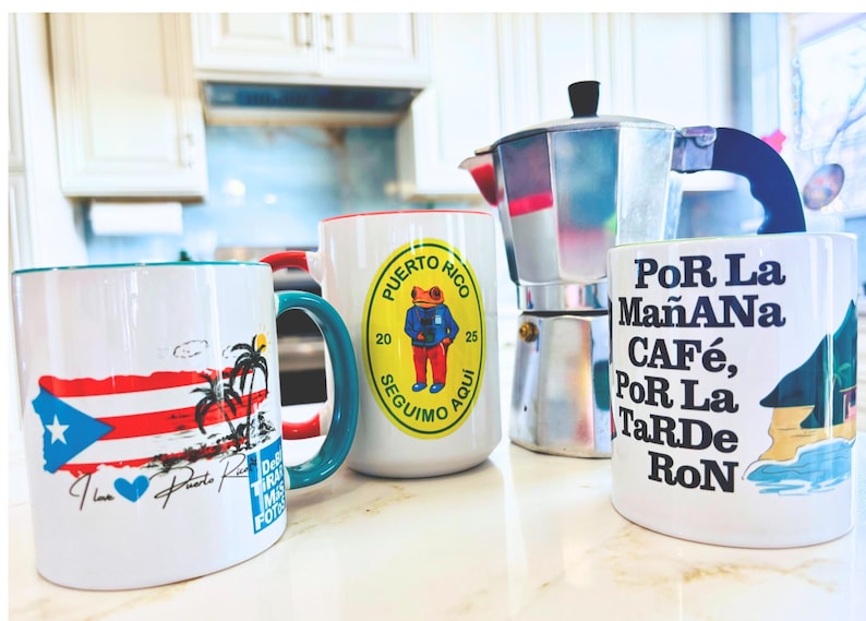 May include: Four white ceramic mugs with different designs. One mug features a Puerto Rican flag, another has a frog wearing a blue shirt, a third has the text "Por La Ma&ntilde;ana Caf&eacute;, Por La Tarde Ron", and the last mug has a palm tree and a beach scene. A silver coffee pot is also in the image.