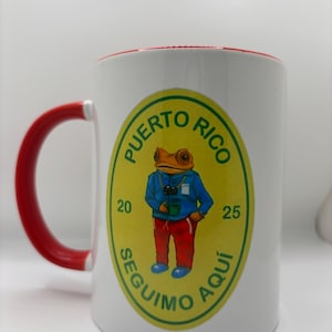 May include: White ceramic mug with a red rim and handle. The mug features a yellow and green circular design with the text "PUERTO RICO" and "SEGUIMO AQUI" around a cartoon frog wearing a blue jacket and red pants.