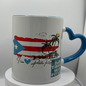May include: White ceramic mug with a blue handle. The mug features a colorful image of the Puerto Rican flag, palm trees, and the text "I love Puerto Rico". The text "Debi Tirar Mas Fotos" is also printed on the mug.