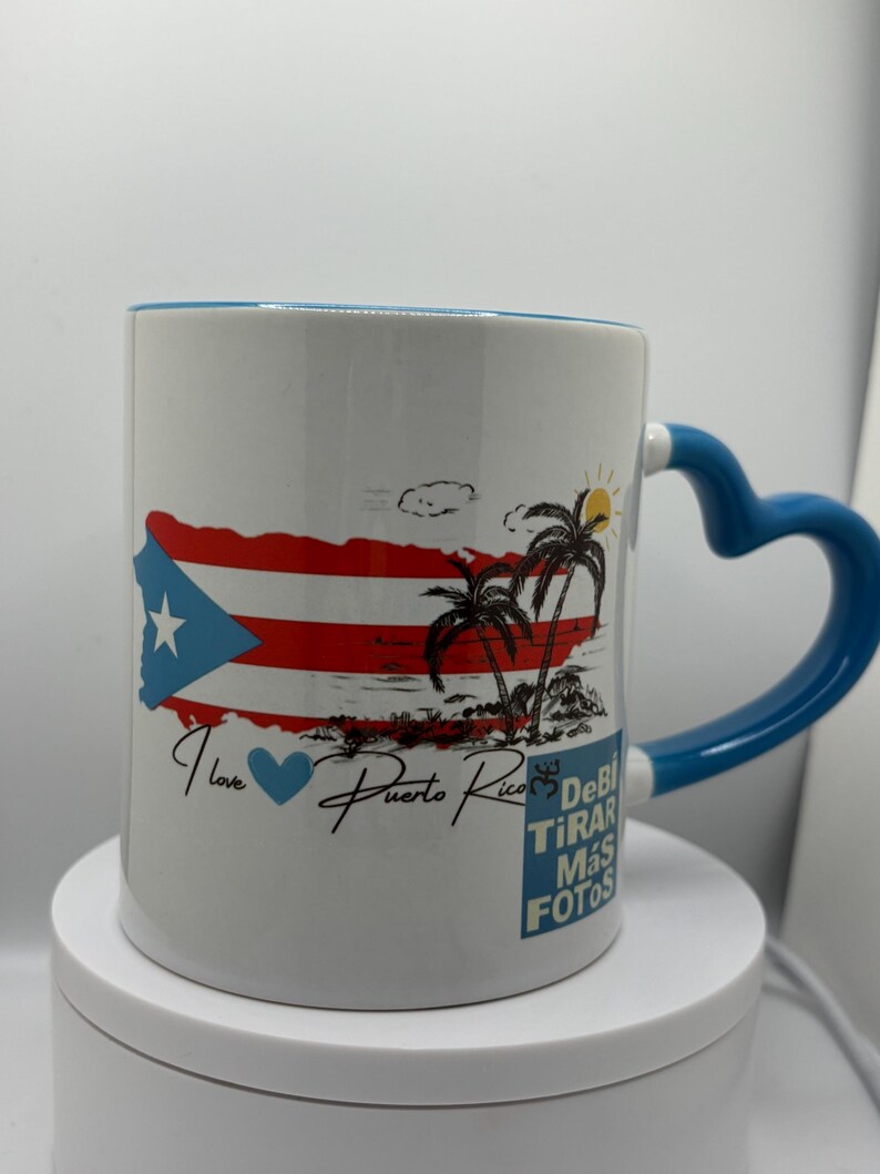 May include: White ceramic mug with a blue handle. The mug features a colorful image of the Puerto Rican flag, palm trees, and the text "I love Puerto Rico". The text "Debi Tirar Mas Fotos" is also printed on the mug.