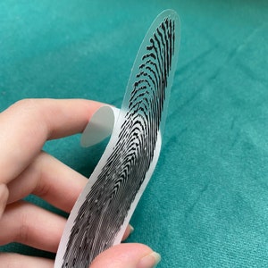 May include: A clear plastic sheet with a black fingerprint design printed on it. The sheet is curved and held by a hand.