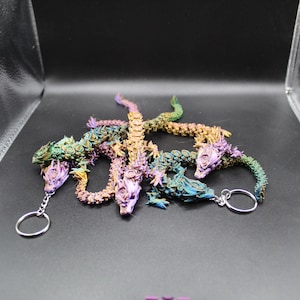 May include: A collection of articulated dragon keychains in various iridescent colors, including purple, green, gold, and blue. Each dragon has a detailed head and body, and is attached to a silver keyring. The keychains are displayed on a dark surface.