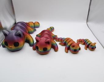May include: Four rainbow-colored cat figurines in different sizes. The largest cat is purple, the next largest is red, and the two smallest cats are orange. The cats are all in a curled-up position.
