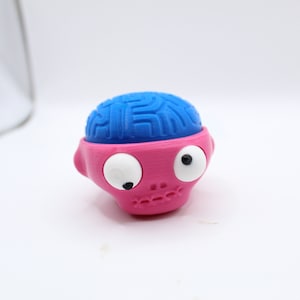 May include: A pink and blue toy with googly eyes and a brain-shaped head. The toy is made of plastic and has a cartoonish appearance.