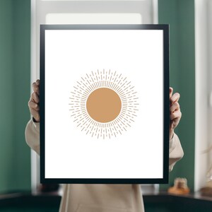 Puede incluir: Una impresión artística enmarcada con un diseño de sol minimalista. El sol se representa con un círculo central de color marrón claro y líneas radiantes en un tono similar sobre un fondo blanco. El marco es negro.