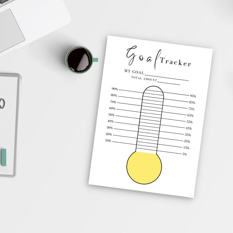 A yellow Goal Tracker is shown here on a white table beside a coffee mug and laptop.