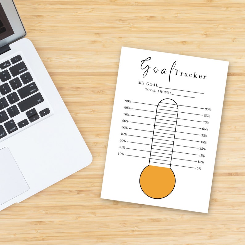 The orange Goal Thermometer on a light, wooden desk beside a laptop.