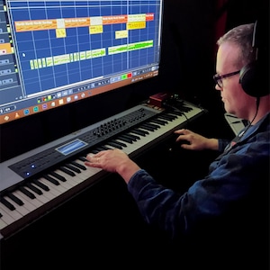 May include: A person sits in front of a computer screen with a music production program open. They are playing a keyboard with their right hand. The screen shows a multi-track audio recording with various colored waveforms.