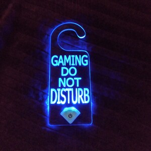 以下が含まれることがあります： 「GAMING DO NOT DISTURB」という文字と電源ボタンが付いた青い照明付きドアハンガーです。
